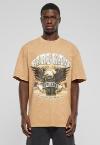 Beige grafisk t-shirt med et ørnedesign og "KARL KANI EST. 1989" trykt med fede bogstaver. Løs pasform og rund halsudskæring.
