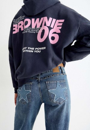 Person, die einen dunklen Hoodie mit pinkem Text und blaue Jeans mit großen Stern-Patches auf den Gesäßtaschen trägt, von hinten gezeigt.