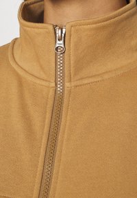 Veste zippée beige avec un col haut et une fermeture éclair en métal. Tissu doux et texturé avec des détails cousus le long des coutures.