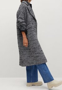 Manteau à motifs pied-de-poule noir et blanc avec larges revers, poches et manches retroussées. Associé à un jean bleu à jambes larges et des chaussures à plateformes beiges.