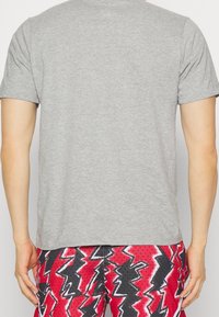 Camiseta de algodón gris con cuello redondo, mangas cortas y un ajuste relajado, mostrada en una persona, combinada con pantalones cortos rojos con estampado.