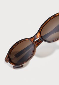Schildpatt-Sonnenbrille mit runden braunen Gläsern, einem glatten Kunststoffrahmen und breiten Bügeln, die ein dezentes Muster und ein glänzendes Finish aufweisen.