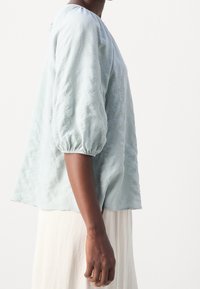 Blouse bleu clair texturée avec des manches bouffantes, un col rond et une coupe décontractée. Le tissu doux donne une apparence détendue.