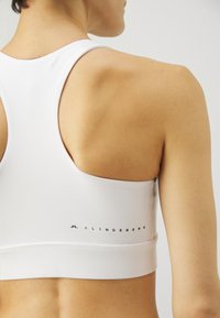 Λευκό αθλητικό σουτιέν με λεία υφή, σχέδιο racerback, με ένα διακριτικό λογότυπο σε μαύρο χρώμα κατά μήκος της κάτω ζώνης.