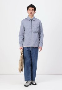 Lys blå, lett jakke med glidelås, brystlomme og rund krage, brukt med blå rette jeans og sølvfargede joggesko.