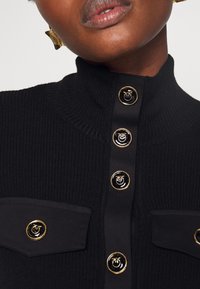 Svart ribbad höghalstop med guldknappar och två fickor med guldaccenter. Slät tygkänsla med en figurnära design.