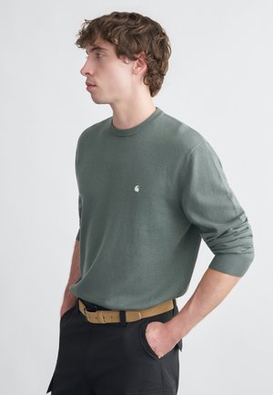 Ανοιχτοπράσινο πλεκτό πουλόβερ με στρογγυλή λαιμόκοψη, ριμπ μανσέτες και μικρό λογότυπο στο στήθος, φορεμένο με μαύρη ζώνη και παντελόνι cargo.