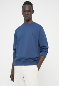 Μπλε βαμβακερό φούτερ με στρογγυλή λαιμόκοψη, ριμπ μανσέτες και τελείωμα. Διαθέτει μικρό λογότυπο στο αριστερό στήθος. Φορεμένο με ανοιχτόχρωμο παντελόνι.