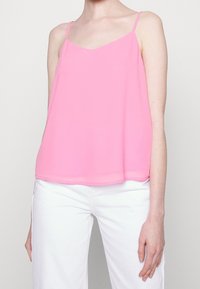 Pink, sleeveless camisole top made of lightweight fabric, featuring a V-neck and straight hem, paired with white pants.
