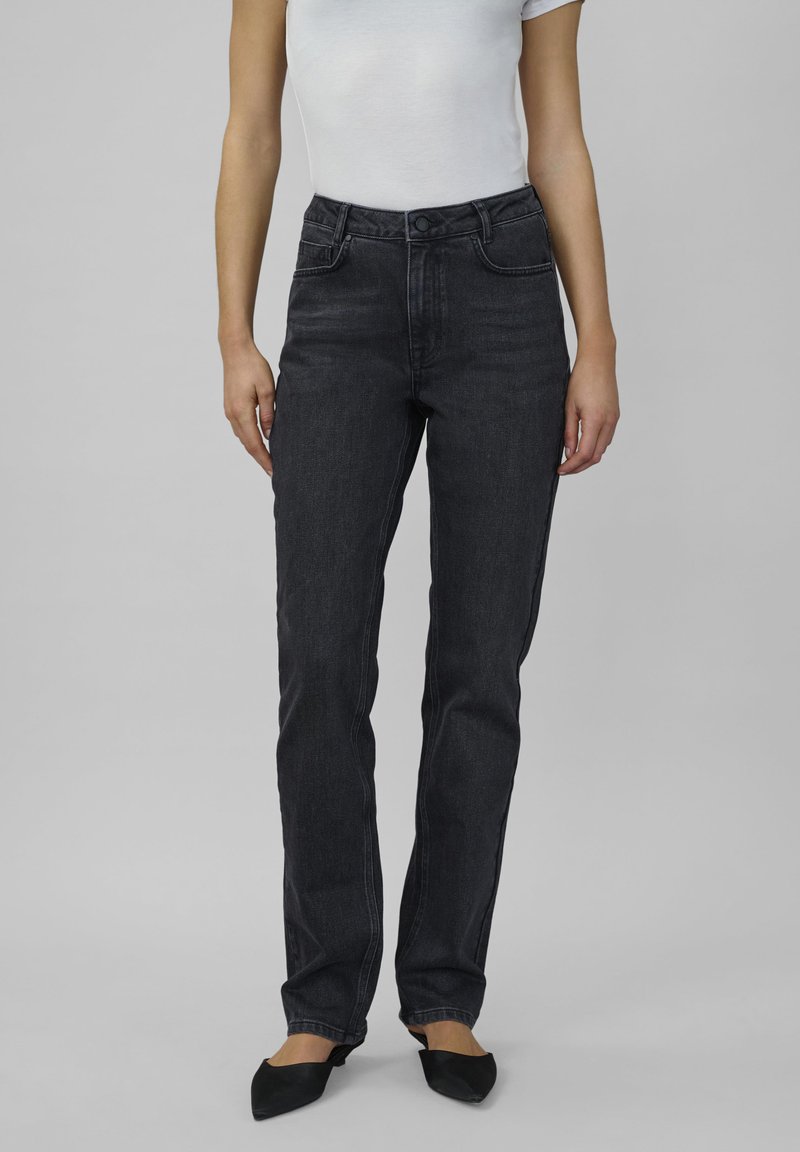 Frau in hoch taillierten schwarzen Jeans und einem weißen, figurbetonten Kurzarmshirt, steht vor einem schlichten grauen Hintergrund.