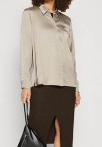 Beige Satinbluse mit einem Knopfkragen, langen Ärmeln und einem seitlichen Schlitz. Kombiniert mit einem gerippten braunen Rock und einer schwarzen Umhängetasche.