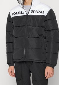 Czarna kurtka puchowa z białym panelem na ramieniu, z logo "KARL" i "KANI". Zamek błyskawiczny i boczne kieszenie, teksturowany wzór pikowania.