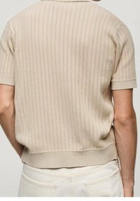 Polo shirt in maglina beige a maniche corte, con una trama a coste, presenta un orlo aderente e un design pulito e semplice. Nessun logo o motivo visibile.