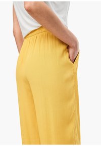 Personne portant un pantalon jaune à taille élastique et une chemise blanche, main placée de manière décontractée dans la poche droite, vue de côté.