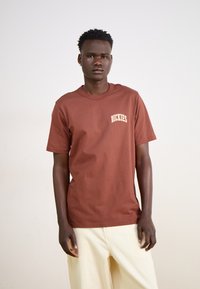 Brūns kokvilnas T-krekls ar īsām piedurknēm un apaļu kakla izgriezumu. Uz kreisā krūtīm ir balts "Dickies" logo. Vienkārša auduma tekstūra.