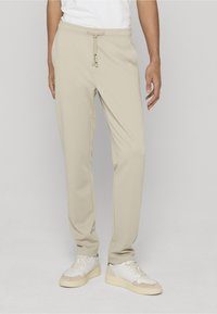 Pantaloni jogger beige con fascia elastica in vita, chiusura con coulisse, tasche laterali e vestibilità comoda, abbinati a sneakers bianche.