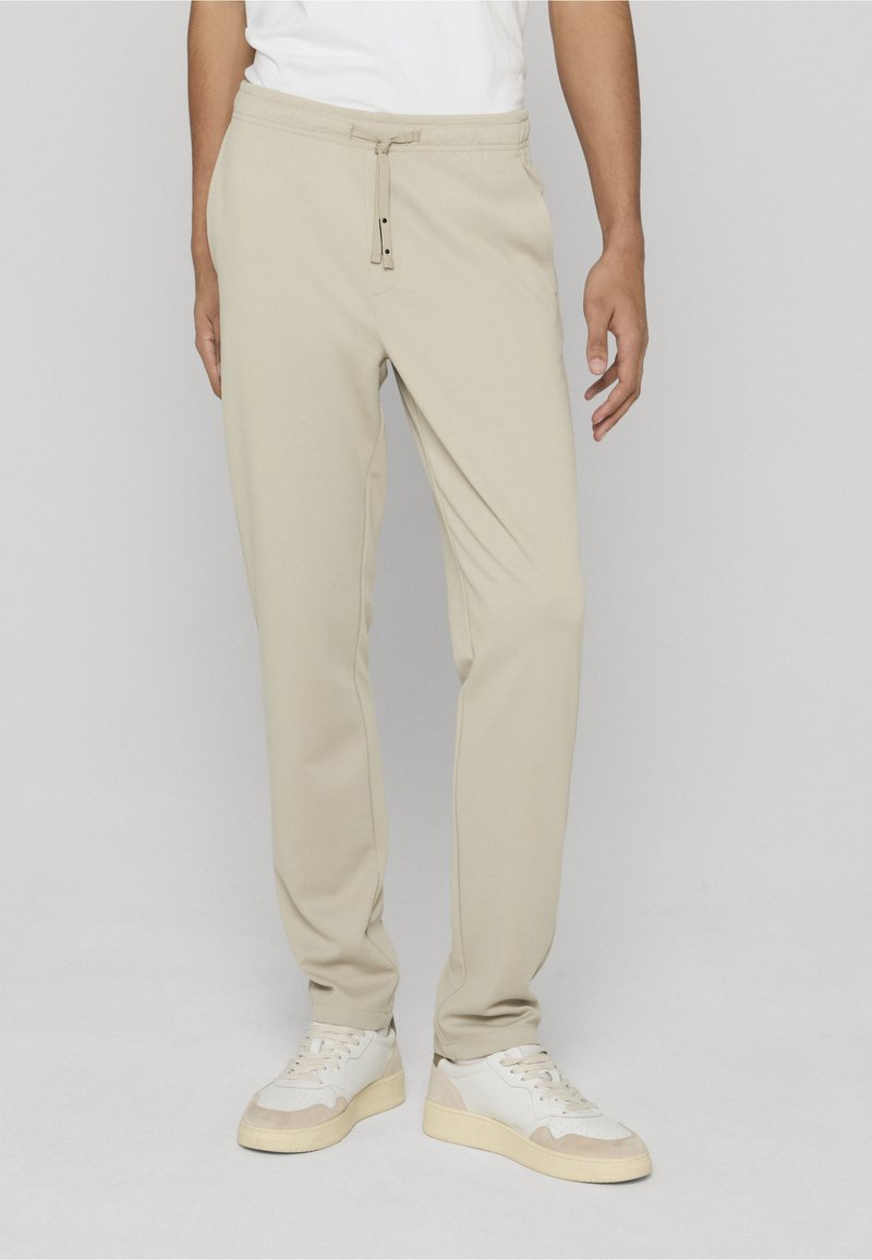 Pantaloni jogger beige con fascia elastica in vita, chiusura con coulisse, tasche laterali e vestibilità comoda, abbinati a sneakers bianche.