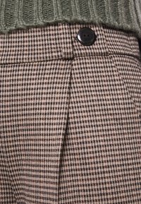 Braun- und cremefarbene karierte Hose mit einem schwarzen Knopf, ausgestattet mit dezenten Falten und einem strukturierten Stoff, kombiniert mit einem grünen gerippten Strickpullover.