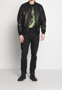 Svart läderbomberjacka med ribbade ärmslut, svart grafisk t-shirt, mörka jeans och svarta snörskor. Enkel, modern design.