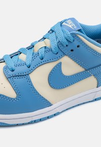Mėlyni ir kreminiai odiniai sportbačiai su tekstūruotu paviršiumi, toniniais siūlais ir raštuotomis aulomis. Yra ryškus mėlynas Nike logotipas.