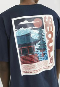 Marineblå bomulds T-shirt med et farverigt grafisk motiv af en traditionel bygning, bjerge og solnedgang, med teksten "SEOUL" i fede bogstaver.