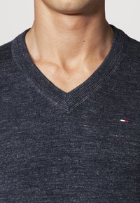 Homme portant un pull gris foncé à col en V avec un petit logo brodé rouge, blanc et bleu sur la poitrine gauche.