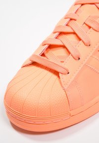 Oranssi lenkkari, jossa on teksturoitu kumikärki, sileä synteettinen päällinen ja litteät nauhat. Sisältää viistosti ommeltuja saumoja ja hienovaraisia kuvioita.
