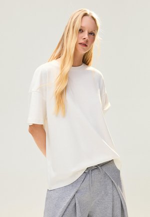 Young woman with long blonde hair wearing a loose white T-shirt and light gray drawstring pants, standing against a plain light background.