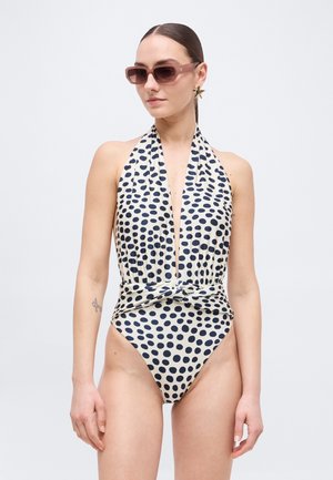Kvinne iført hvit halter-badedrakt med svarte polkadotter, knutedetalj i midjen, stjerneformet øredobb og brune solbriller.