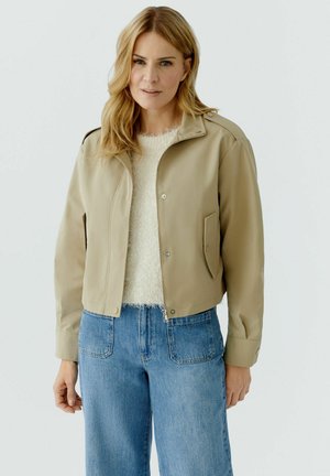 Frau mit blonden Haaren, die eine beige Jacke über einem weißen, strukturierten Pullover und blauen Jeans trägt, steht vor einem schlichten Hintergrund.