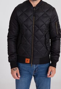 Veste bomber matelassée noire avec poignets et ourlet côtelés. Comprend une fermeture éclair à l'avant, des poches latérales et une étiquette de marque orange en bas.
