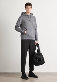 Šedá mikina s kapucí a přední kapsou, doplněná černými zúženými kalhotami a černými botami. Drží černou tašku. Minimalistický design.