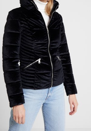 Kvinde iført en sort quiltet fløjlsjakke med sølv lynlåse, hvid rullekrave og lyseblå jeans mod en ensfarvet baggrund.