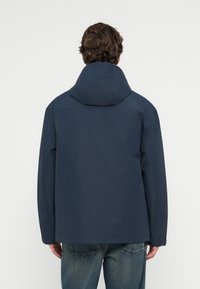 Ναυτικό μπλε μπουφάν με κουκούλα σε χαλαρή εφαρμογή, εντελώς λείο υλικό και ελάχιστες ραφές, συνδυασμένο με ξεθωριασμένο μπλε τζιν.