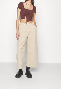 Femme portant un crop top marron froncé, un pantalon beige à jambes larges, des bottes noires épaisses et un collier argenté superposé, debout avec une main dans la poche.