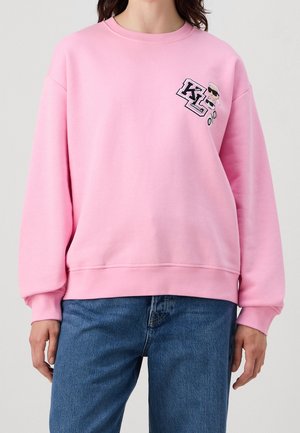 Vrouw draagt een lichtroze sweatshirt met het Karl Lagerfeld-logo en cartoon linksboven, gecombineerd met een blauwe spijkerbroek.