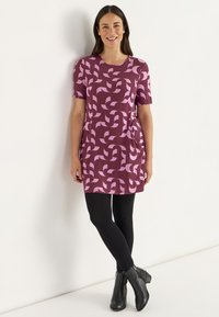 Kurzärmeliges lilanes Kleid mit pinkem abstraktem Muster, ausgestattet mit seitlichen Taschen und einem lässigen Schnitt, kombiniert mit schwarzen Leggings und Ankle Boots.