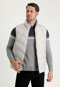 Chaleco acolchado gris claro con cuello alto, textura acolchada, cierre de cremallera y bolsillos laterales, superpuesto a un suéter oscuro azul y gris.