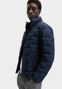 Chaqueta de plumas azul marino con cuello alto y frente con cremallera, que presenta secciones acolchadas y dos bolsillos laterales, llevada sobre pantalones de chándal grises.