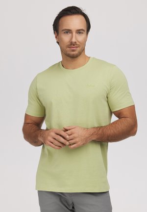 Mężczyzna z krótkimi ciemnymi włosami i lekkim zarostem, ubrany w jasnozieloną koszulę z krótkim rękawem i szare spodnie, stoi na jednolitym tle.