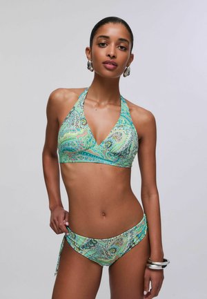 Donna che indossa un bikini halter verde con motivo paisley, orecchini a cerchio d'argento e bracciali, in posa su uno sfondo grigio uniforme.