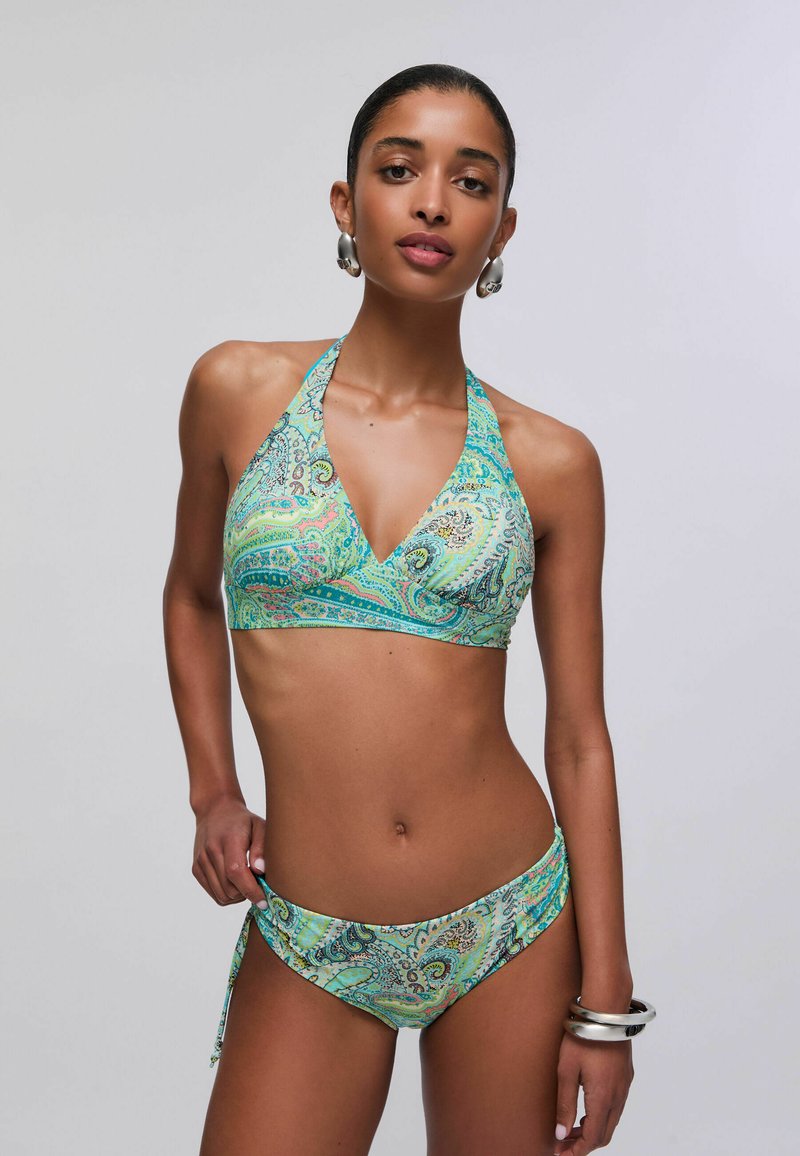 Femme portant un bikini halter vert à motif cachemire avec des boucles d'oreilles créoles argentées et des bracelets, posant devant un fond gris uni.
