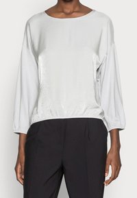 Light gray, shiny long-sleeve top with a wide neckline and elastic hem, paired with black pants. Soft texture, smooth finish, relaxed fit.