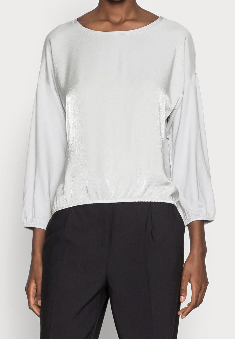 Light gray, shiny long-sleeve top with a wide neckline and elastic hem, paired with black pants. Soft texture, smooth finish, relaxed fit.