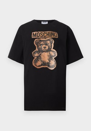 Naine seisab ja kannab lahtist musta T-särki, millel on pruun kaisuloom ja tekst "MOSCHINO", hele siniste teksapükste, mustade kontsadega, käed taskutes.