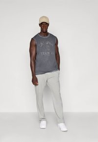 T-shirt cinzento sem mangas com estampa, calças de fato de treino cinza claro e ténis brancos. O modelo usa um boné bege. Texturas de tecido suaves e casuais.