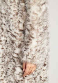 Płaszcz z futerka sztucznego w jasnobeżowym kolorze z ciemnymi, cętkowanymi wzorami. Charakteryzuje się długą, miękką fakturą i przylegającym fasonem przy rękawach. Ręka widoczna z boku.