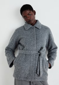 Grauer Wollmantel mit Bindegürtel, ausgestattet mit einem kragenförmigen Ausschnitt, Knopfverschluss und strukturiertem Strickdesign. Die Taschen sind in die Seitennaht eingearbeitet.