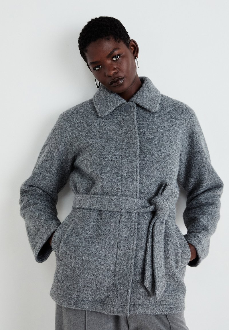 Grauer Wollmantel mit Bindegürtel, ausgestattet mit einem kragenförmigen Ausschnitt, Knopfverschluss und strukturiertem Strickdesign. Die Taschen sind in die Seitennaht eingearbeitet.