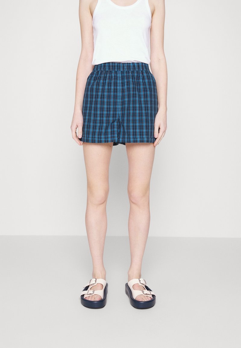 Blå rutiga shorts med elastisk midja, ihop med en vit tanktopp och tvåfärgade sandaler med justerbara spännen.